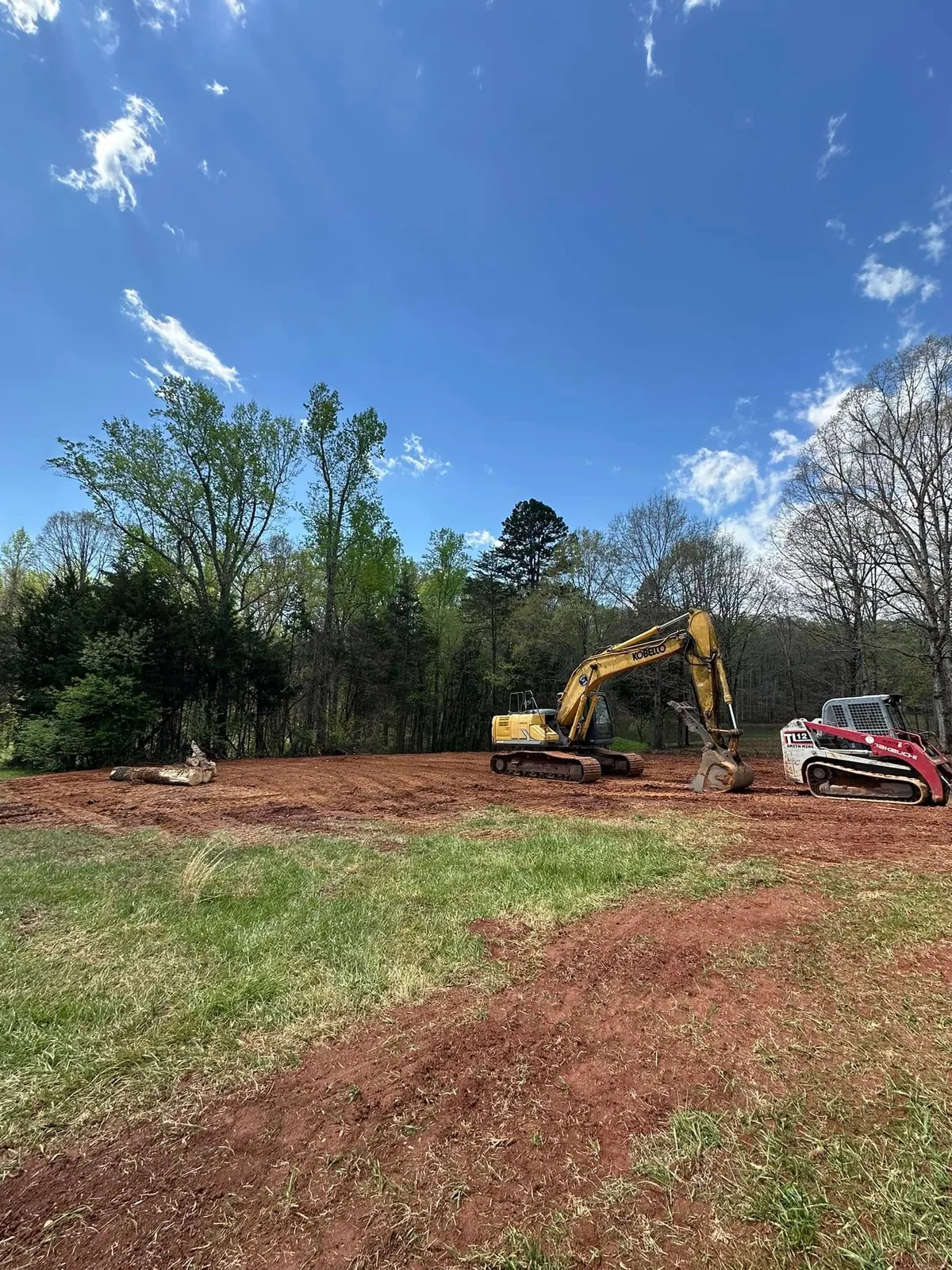Kobelco excavator and Takeuchi TL12 track loader on freshly cleared red-dirt lot in spring