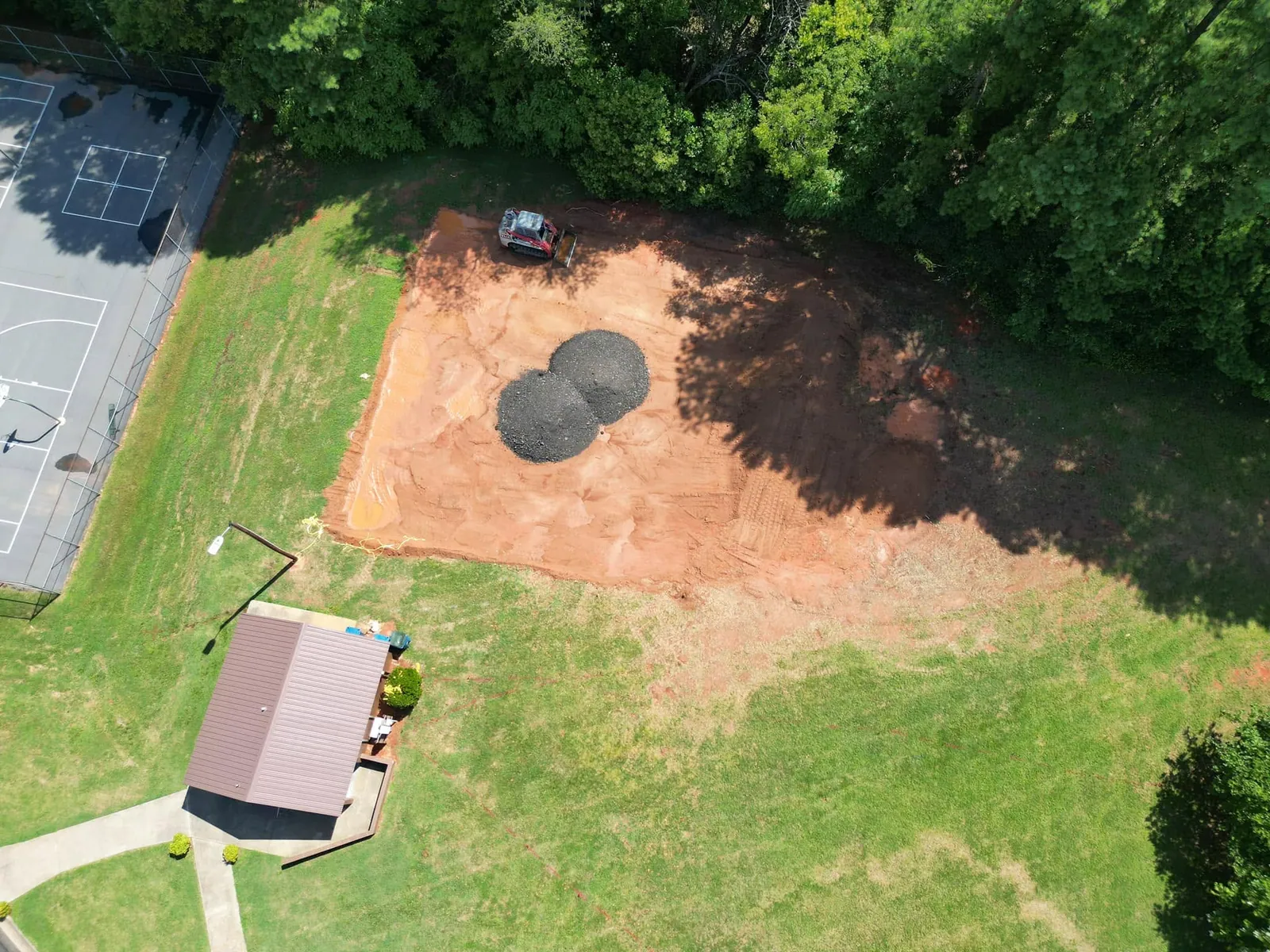 Aerial view of red-clay pad graded beside tennis courts, track loader and two gravel piles in Claremont, NC