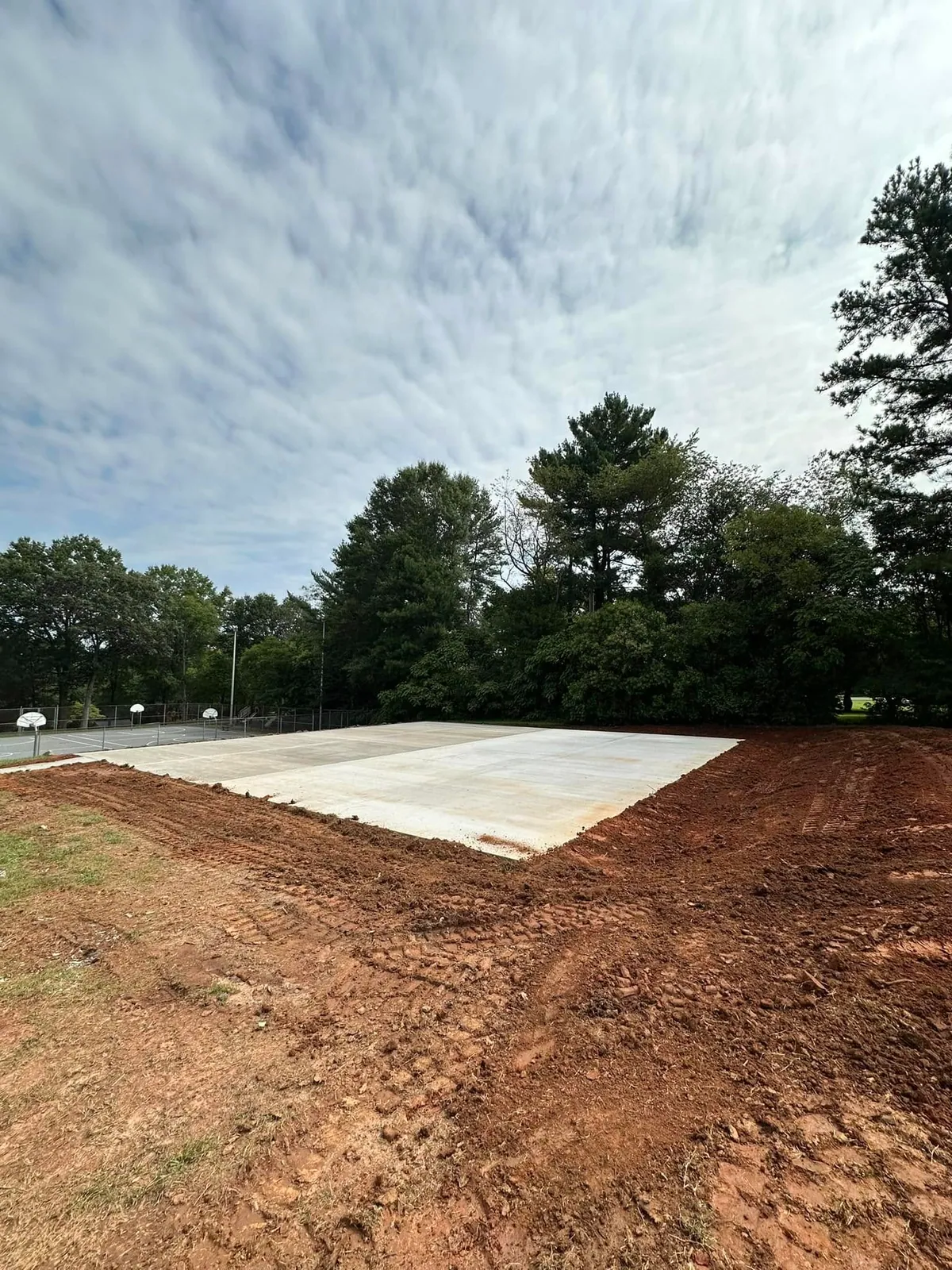 Fresh concrete pickleball court slab poured at park beside basketball court, red clay graded around in Claremont, NC