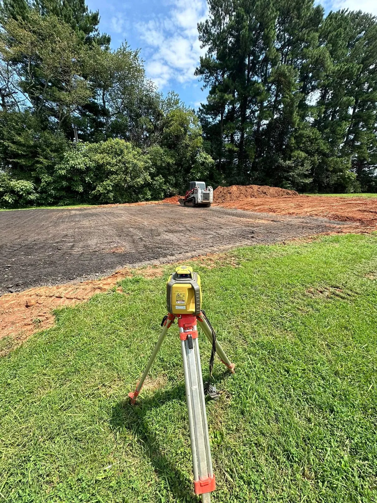 Laser level on tripod; Takeuchi track loader spreading crushed stone base on court pad in Claremont, NC