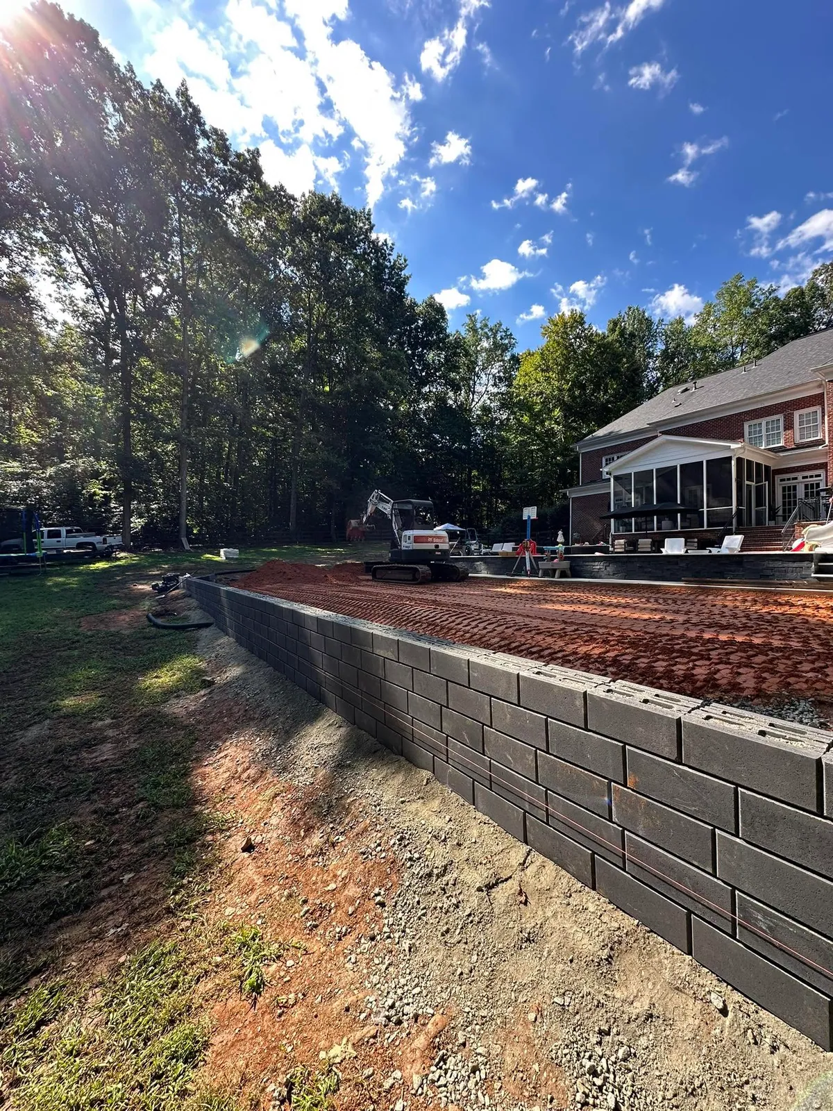 Bobcat mini excavator grading red clay pad above new block retaining wall behind large brick home