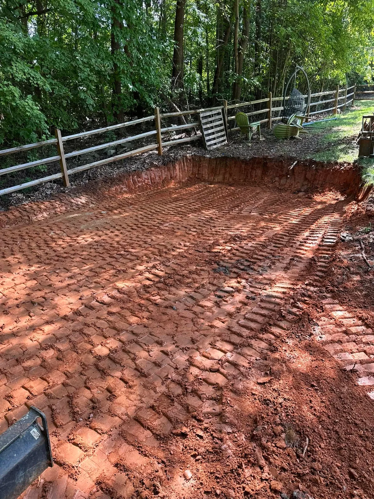 Excavated red clay bench with vertical cut face for retaining wall and swing set, split-rail fence