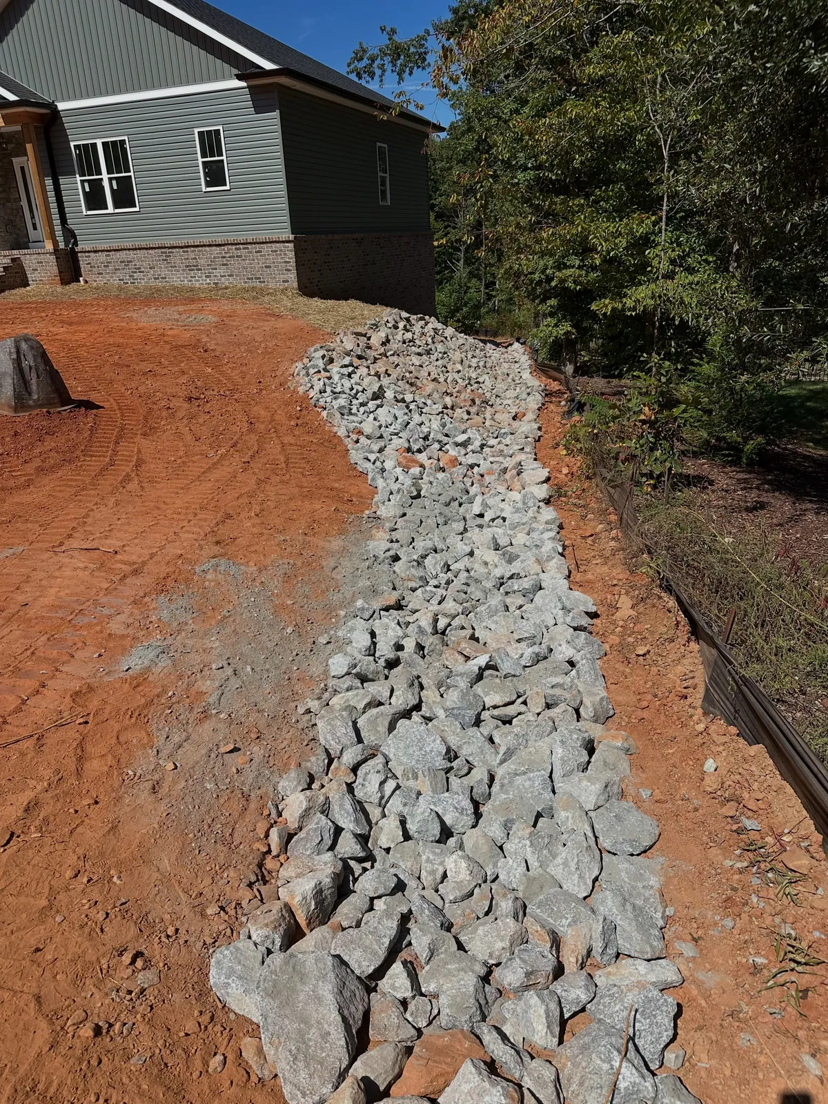 Rip-rap drainage channel running downslope beside new gray house, graded red clay, silt fence