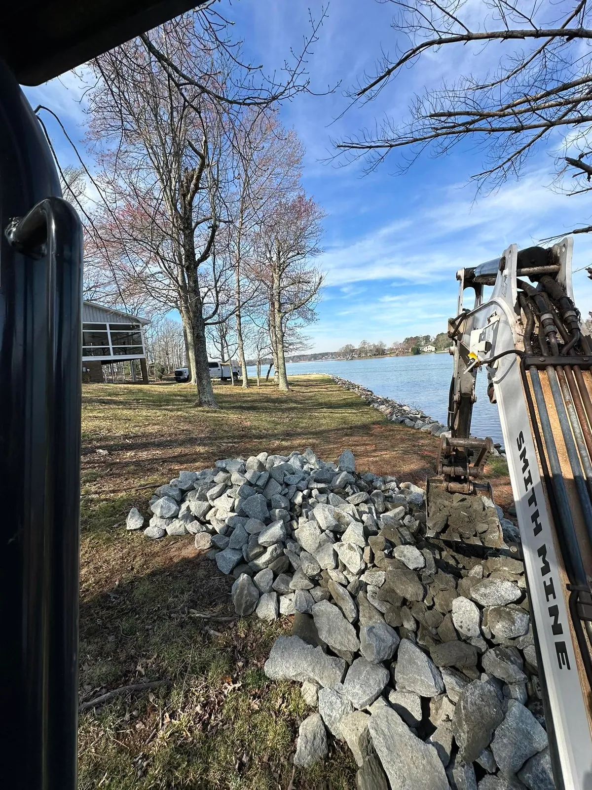 Excavator cab view of rip-rap stone pile on lakefront lawn; existing shoreline riprap and lake behind