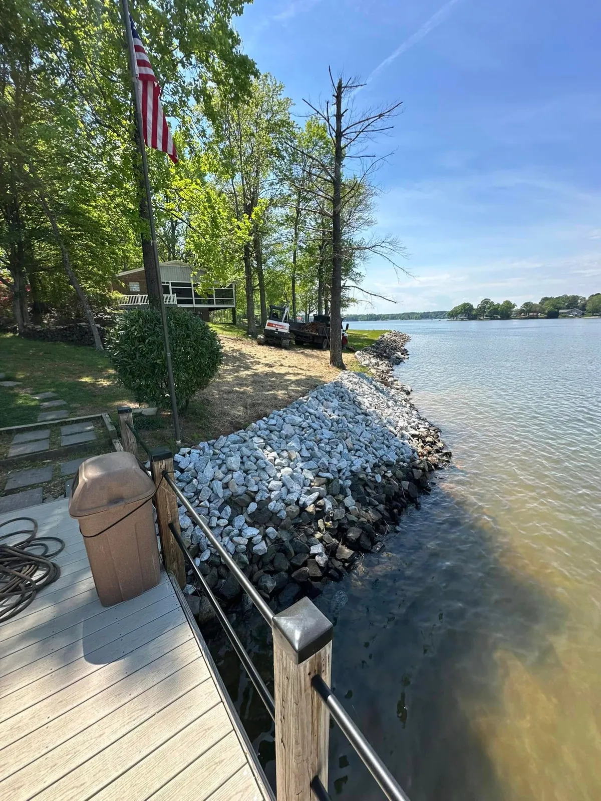 Finished rip-rap stone shoreline along lake beside dock; Bobcat mini excavator and dump trailer on lawn behind