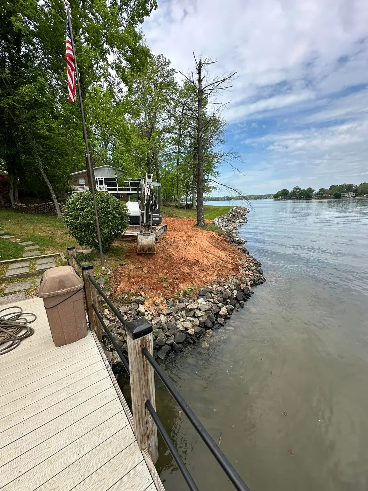 Bobcat mini excavator placing rip-rap along lake shoreline beside dock, American flag, lake house behind