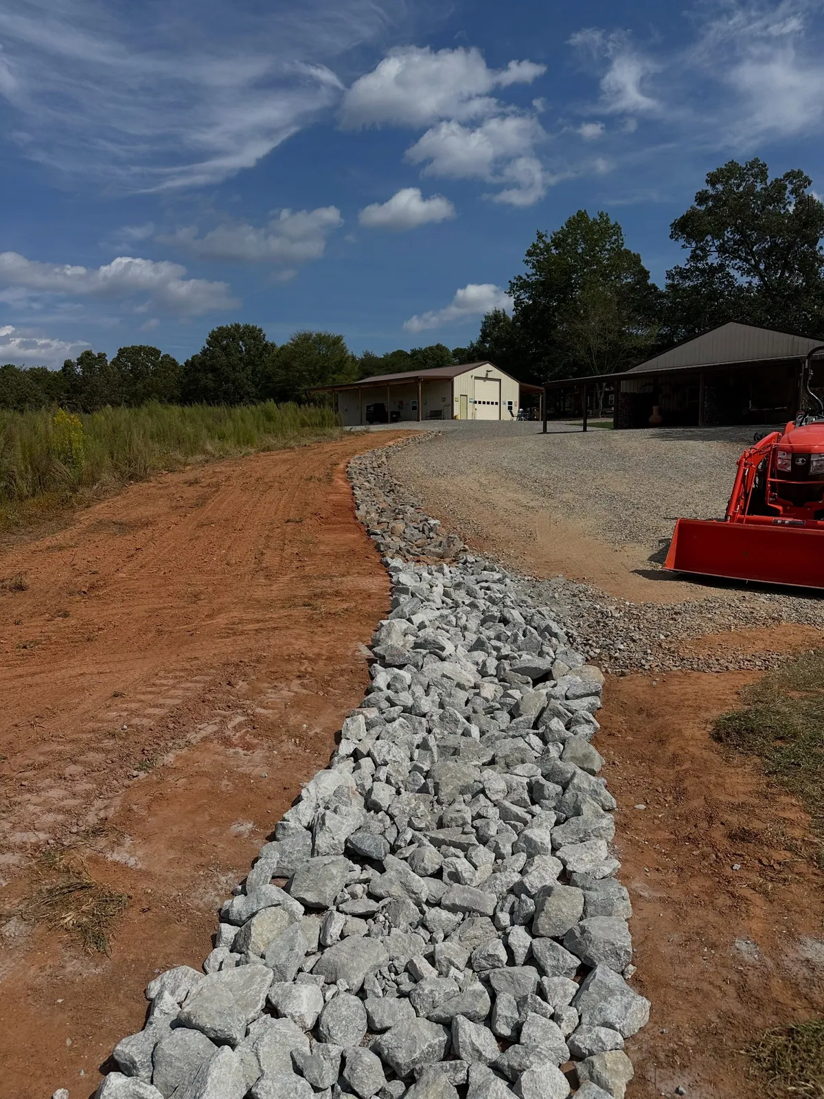 Rip-rap lined drainage swale along graded red clay driveway edge toward metal shop, Kubota tractor