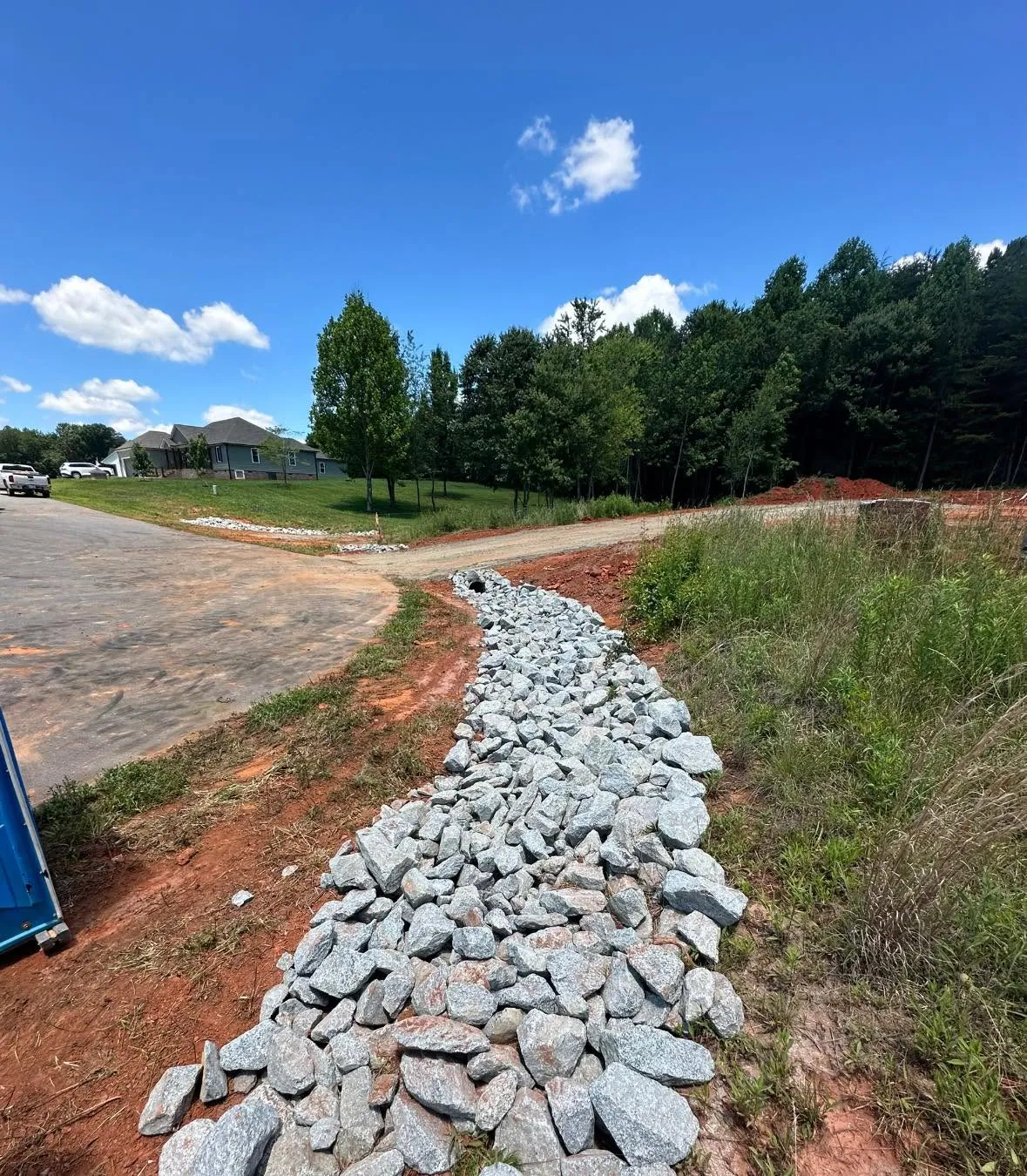 Finished rip-rap lined ditch running from gravel driveway culvert toward road; neighboring house across street