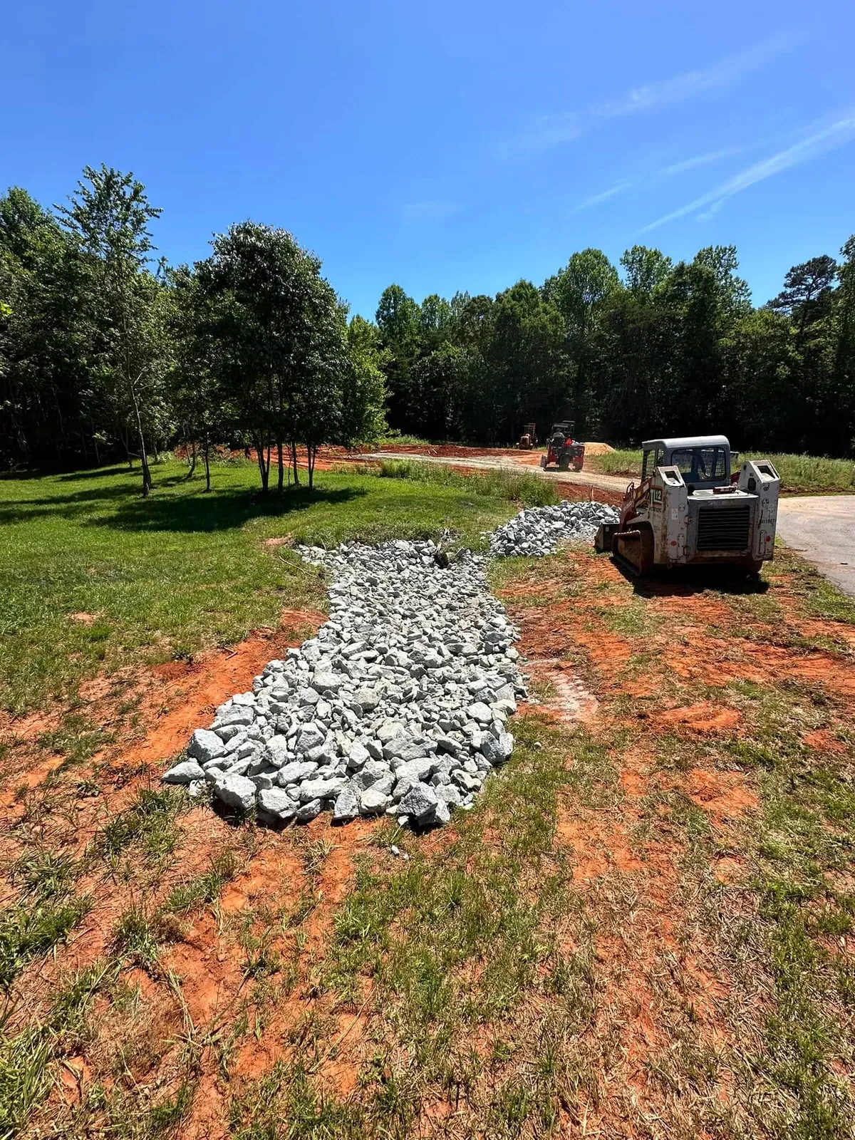 Rip-rap stone drainage swale being laid across grassy yard; Takeuchi TL12 track loader parked beside it