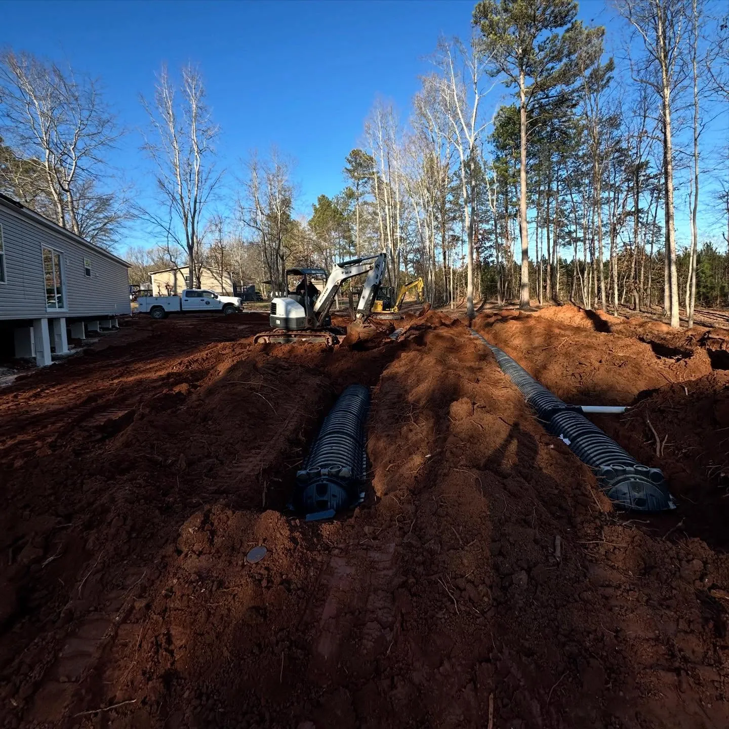 Septic drain field: two Infiltrator chamber runs in red clay trenches beside new house, mini excavators behind