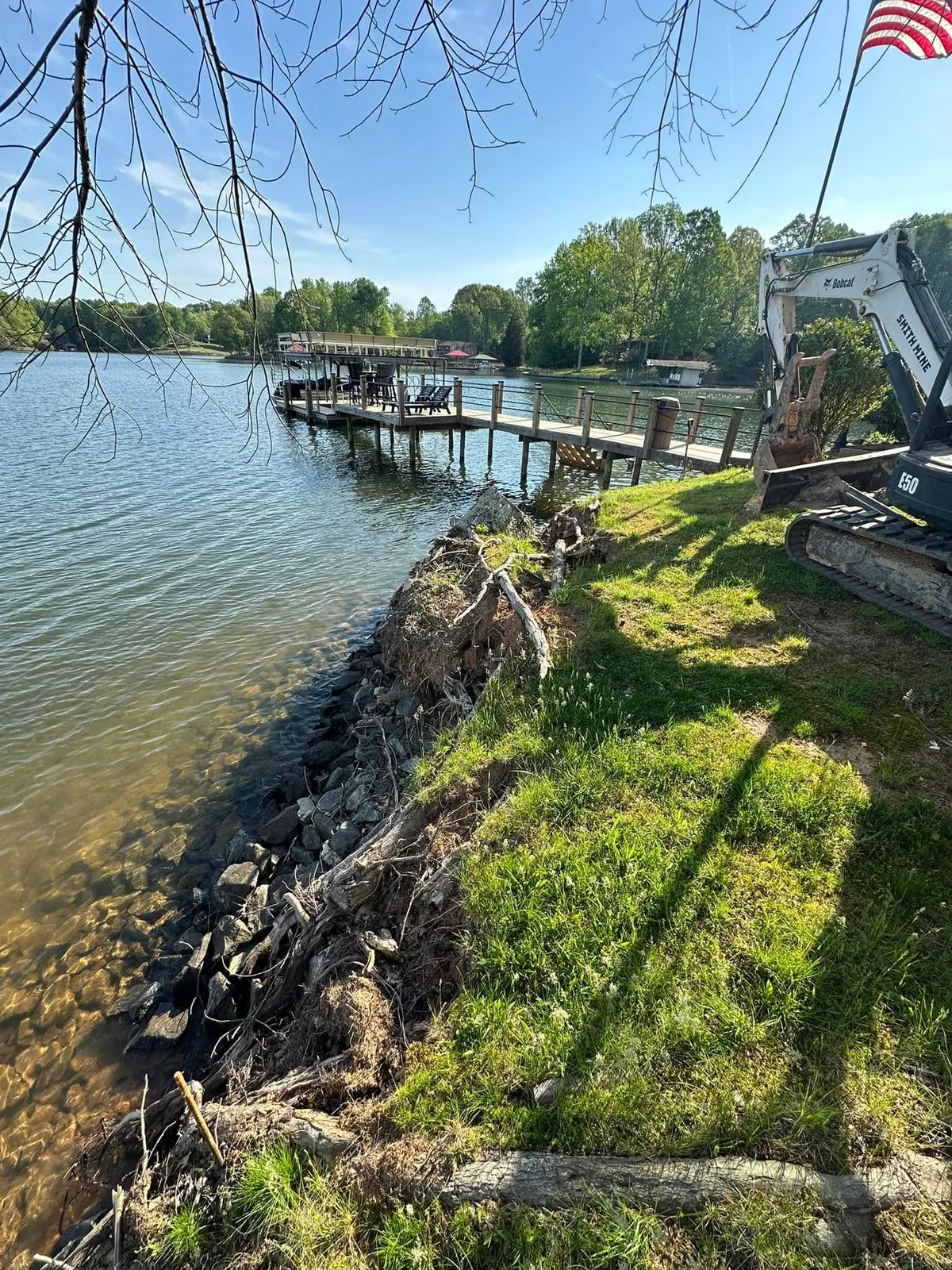 Eroded lake bank with exposed roots before rip-rap; Smith Mine Bobcat E50 mini excavator staged on lawn