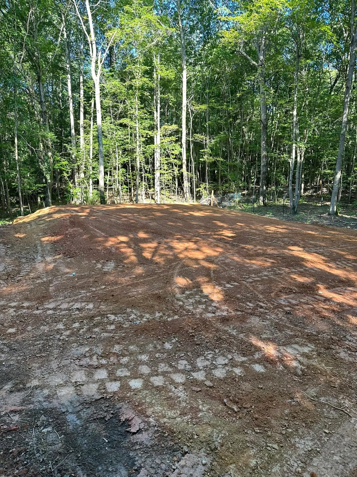 Graded, track-compacted red clay pad in cleared woods, welded-wire fence at tree line behind