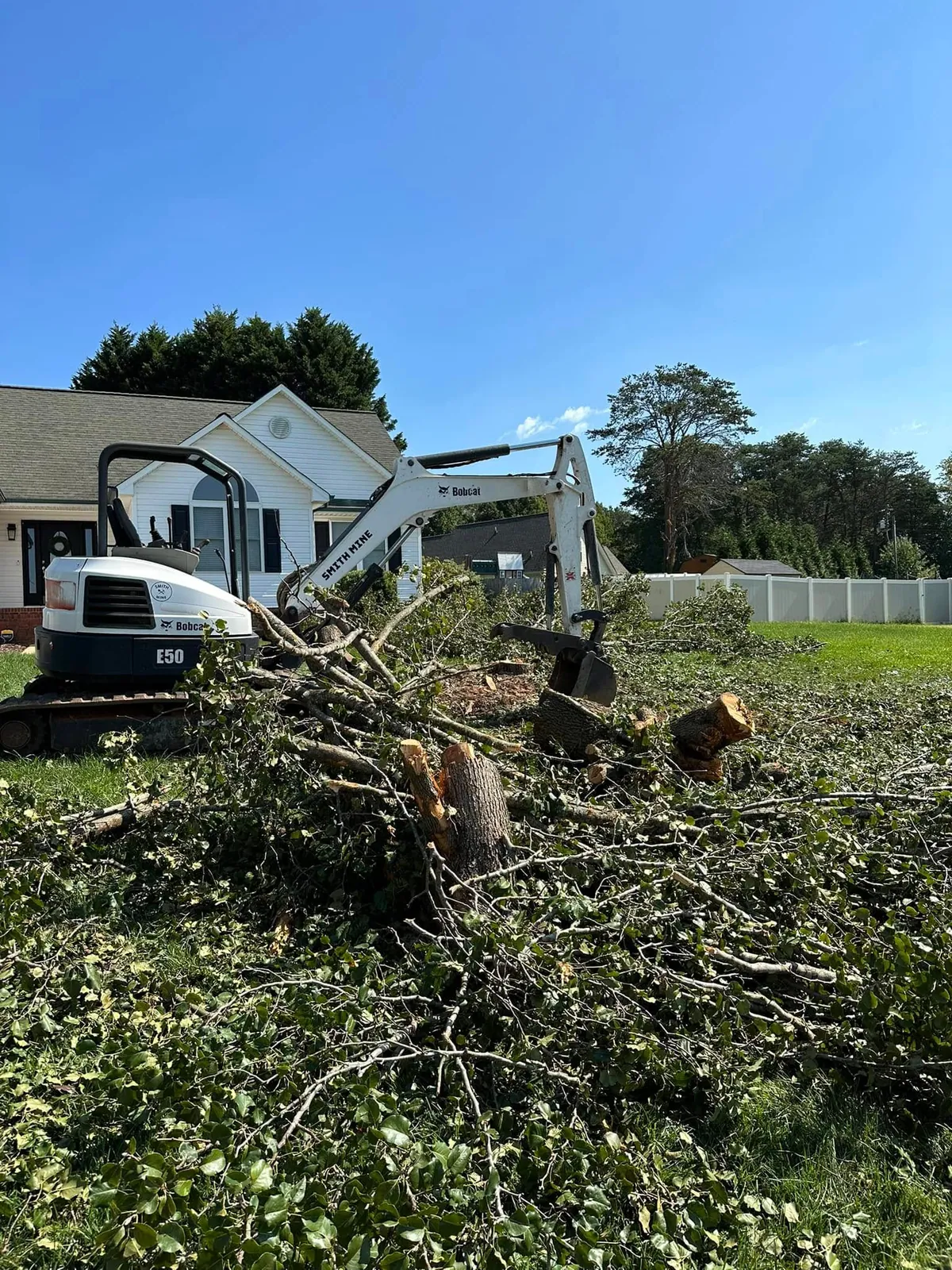 Bobcat E50 mini excavator dragging cut Bradford pear limbs across front lawn of white vinyl house