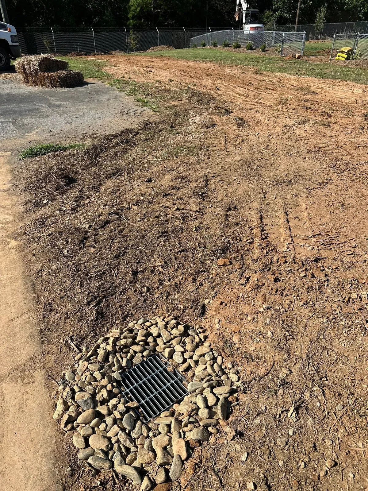 Finished catch-basin grate ringed with river rock on graded strip; mini excavator and truck at fence line