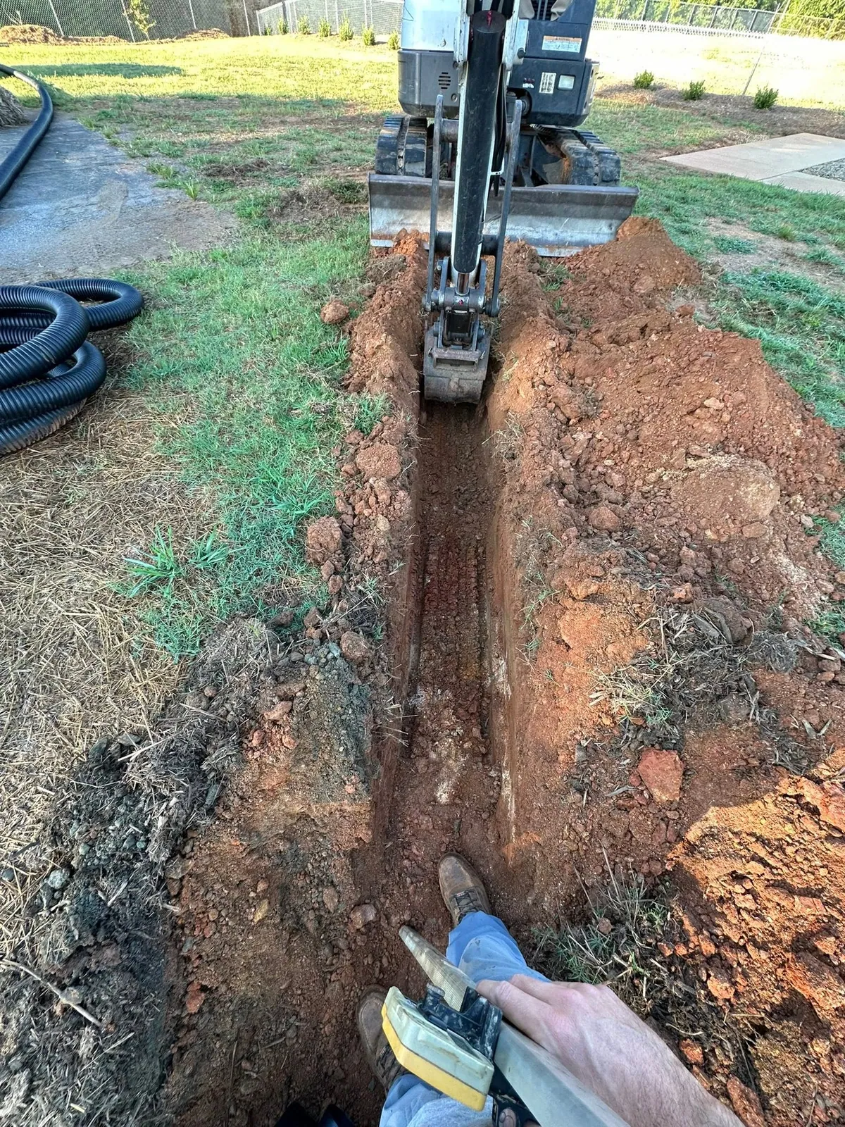 Mini excavator trenching narrow drain line through red clay lawn; black corrugated pipe coiled beside trench